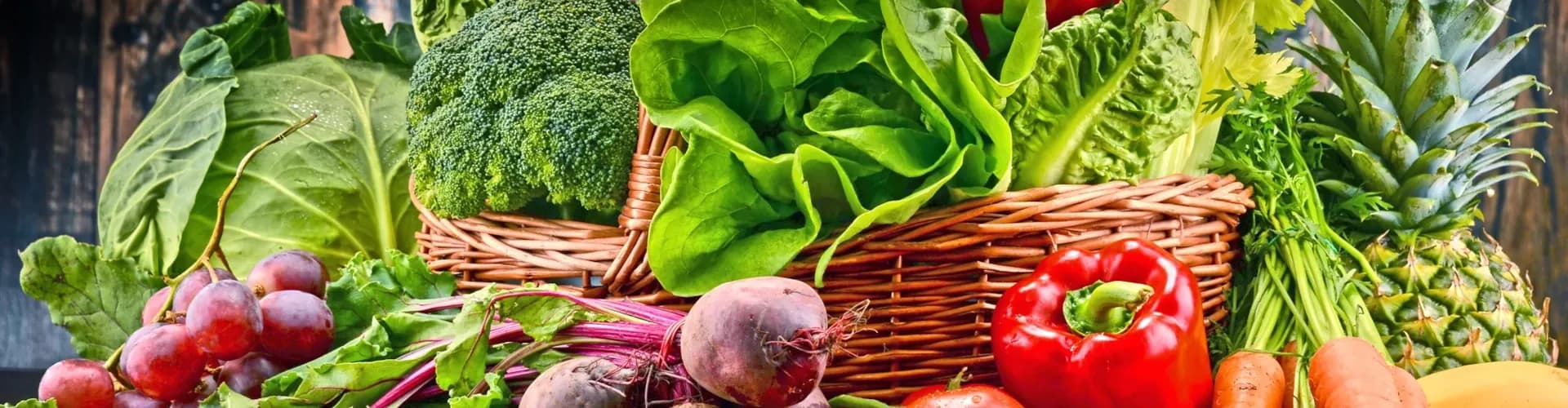 Produce basket filled and surounded by tomatoes, grapes, beets, carrots, pineapples, lettuce, cabbage, bell peppers, and broccoli